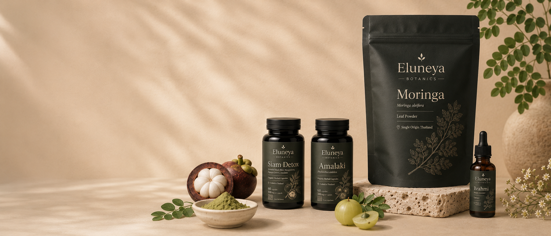 Eluneya Botanics collection — a large Moringa leaf-powder pouch with smaller Siam Detox and Amalaki capsule jars and a small Brahmi dropper bottle, arranged with a bowl of powder and fresh botanicals on a warm beige surface